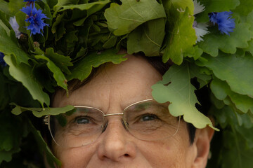 Close-up of an elderly woman wearing a lush oak leaf wreath adorned with blue cornflowers,...