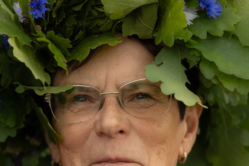 Close-up of an elderly woman wearing a lush oak leaf wreath adorned with blue cornflowers,...