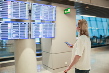 A woman in an airport terminal checks her flight on the departures screen while standing beside her...
