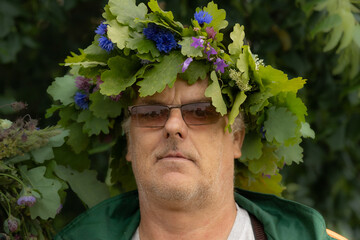 Close-up of a mature man wearing a festive oak leaf and wildflower crown, sunglasses, and a green...