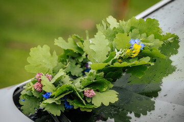 A handmade floral wreath of oak leaves and wildflowers lies on the windshield of a white car,...
