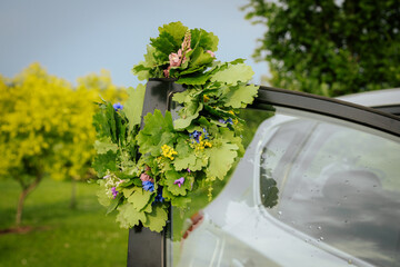 A bouquet of fresh oak leaves and wildflowers decorates the top corner of a car window, symbolizing...