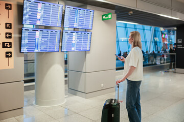 A young woman stands in an airport terminal with her suitcase, looking at the flight departure...