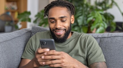 A young man with a beard and dreadlocks smiles while looking at his smartphone on a gray sofa