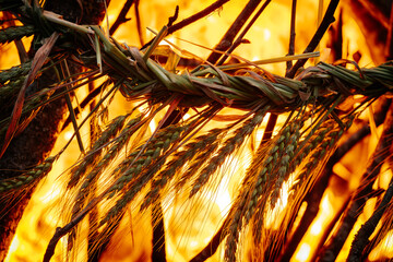 Close-up of a traditional braided wheat ornament burning in vivid orange flames, surrounded by...