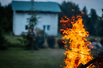 Bright orange flames rise from a bonfire in the foreground, with a blurred house and dark trees in...