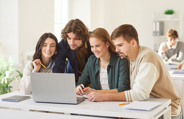 Group of young smiling college or university students sitting at the desk in classroom and talking during a lesson discussing educational project on laptop. Education and knowledge concept.