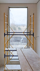 Renovation scene showing a flight of stairs beside a large, dusty window with scaffolding installed for construction work. The space is unfinished, highlighting ongoing interior remodeling efforts.