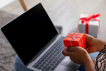 Young black woman using laptop with blank screen, holding gift box, over shoulder shot, closeup....
