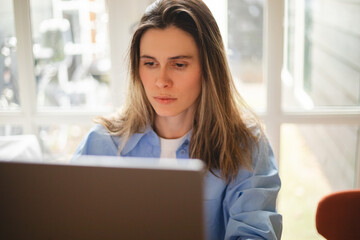 Focused woman freelancer working online on laptop, sitting at desk at home, look at screen, typing, serious young female writing blog or chatting with friends in social network. Girl wear blue shirt.