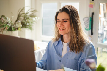 Beautiful young  woman working using computer laptop concentrated and smiling, she sitting at home, freelancer, girl have video call conference. Woman wear blue shirt, look happy.