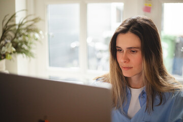 Focused woman freelancer working online on laptop, sitting at desk at home, look at screen, typing, serious young female writing blog or chatting with friends in social network. Girl wear blue shirt.
