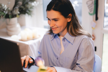 Smiling young woman sit at desk work on laptop or watching webinar, happy girl feel excited browsing internet on computer, using wireless home connection. Technology concept, girl wear blue shirt.
