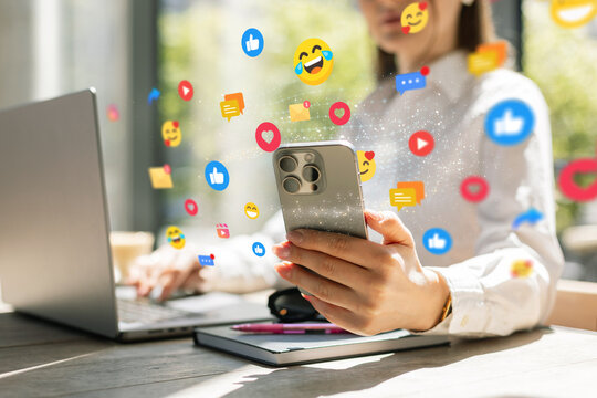 Woman using smartphone with social media icons floating around, sitting at desk with laptop. Concept of digital communication, online interaction, and modern technology in everyday life.
