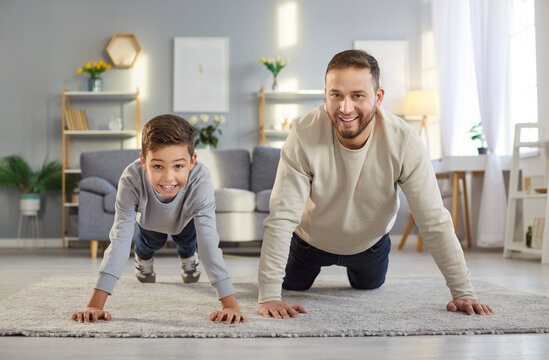 Front view of happy father and son doing push ups together on carpet in living room. Smiling dad and child enjoying home workout, bonding through exercise, encouraging healthy and active lifestyle.