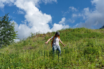Young woman hiker in cap walking on a grassy hillside under a bright blue sky with fluffy clouds, enjoying nature and the outdoors while exploring the natural landscape