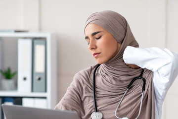 Exhausted muslim woman in hijab doctor rubbing her neck while working with laptop in her cabinet in...