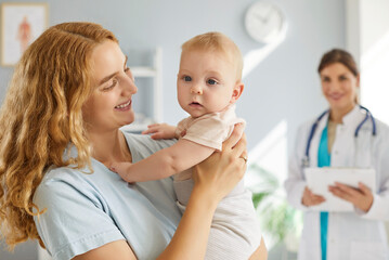 Fototapeta premium Portrait of happy smiling young mother with infant toddler cute little child baby visiting female friendly pediatrician doctor for medical examination standing in clinic. Health care concept.