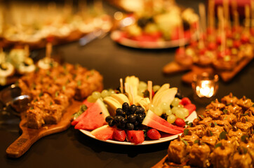Assorted fresh fruits and bite-sized desserts beautifully arranged on a catering table. Warm lighting with a candle adds a cozy and elegant atmosphere for festive events.