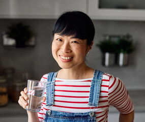 Portrait of a young happy asian woman drinking water and standing in the kitchen