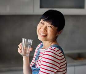 Portrait of a young happy asian woman drinking water and standing in the kitchen