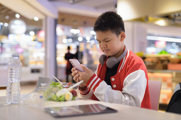 Fototapeta premium Teen Thai boy wearing headphones eating salad in cafeteria texting with phone