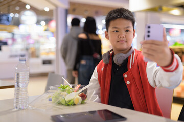 Teen Thai boy wearing headphones eating salad in cafeteria taking selfie with phone