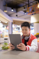 Teen Thai boy wearing headphones eating salad in cafeteria with phone