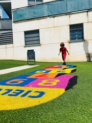 Young boy in red clothes playing hopscotch on a vibrant play area in a sunny urban courtyard.