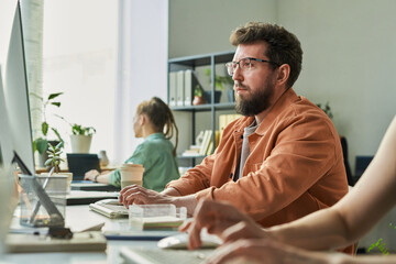 Caucasian young adult man with beard working on neural network development at computer in modern office, sitting at desk with focused expression, colleagues collaborating in background