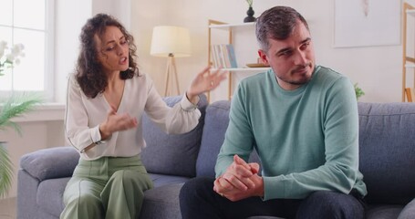 Couple is arguing. During family conflict, irritated husband is tired of his wife scolding or lecturing him. Millennial Caucasian woman passionately tells something to man sitting on sofa at home. - Powered by Adobe