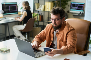 Caucasian middle aged man developing neural network using laptop and smartphone at desk, young...