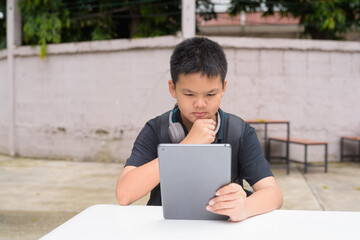 Thai teenage boy sitting at outdoor table using tablet wearing headphones