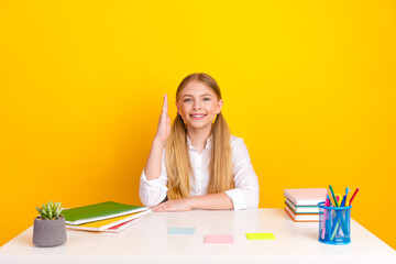 Smiling schoolgirl in white shirt sitting at table with notebooks and raised hand on yellow...
