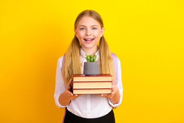 Young schoolgirl with a stack of books and a small plant, standing against a bright yellow...
