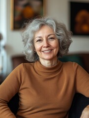 An elderly woman with gray hair, wearing a brown sweater, sits comfortably in her home's living room. She is smiling and appears relaxed as if engaged in friendly conversation.