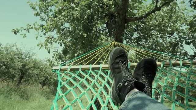 Point-of-view shot of a person lying in a hammock, wearing hiking boots and jeans, gently swinging with a view of trees above. Captures the essence of outdoor relaxation, slow travel, and mindful livi