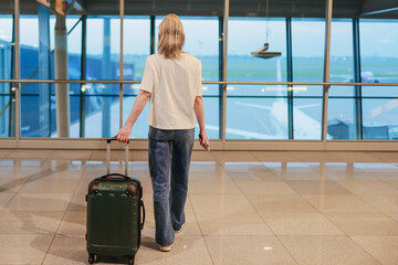 Full-length view of a woman with a suitcase looking out the airport window at airplanes on the...