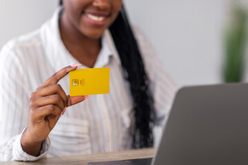Credit card in cheerful black lady hand, pretty african american young woman in official outfit using laptop at home, having remote job, freelancer using online banking or shopping online, empty space