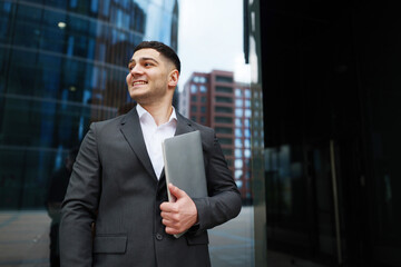 Business professional exudes confidence while holding a laptop outside modern office buildings in...