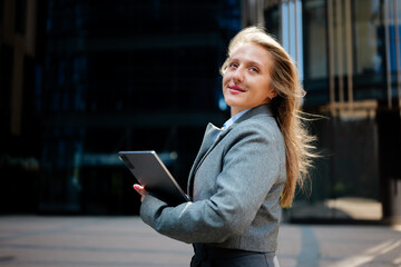 Professional woman smiling confidently while holding a tablet outside a modern building on a sunny...