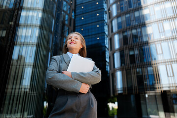 Businesswoman confidently posing with documents in a modern urban setting under gleaming...