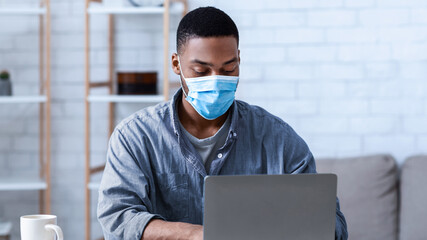 Black Man Wearing Face Mask Working On Laptop Sitting In Modern Office. Social Distancing At Work...