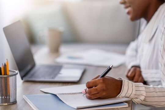 Closeup of young black woman having online course, smiling african american lady in headset sitting at table in front of laptop, taking notes in notepad, home interior, side view, copy space