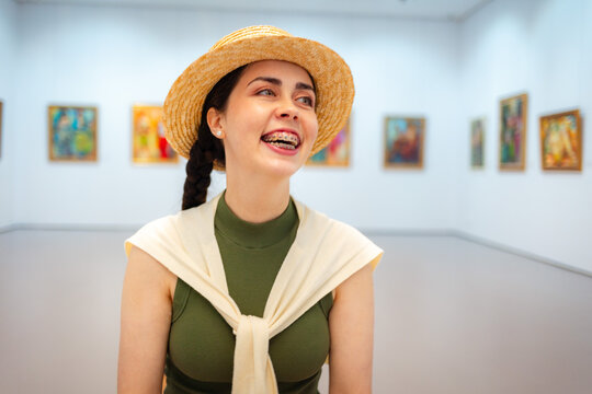 Portrait of young Caucasian happy elegant woman in straw hat smiling in the modern art gallery. Concept of cultural education and Museum Day