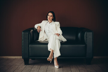 Confident businesswoman in a stylish white suit sitting on a black leather sofa against a brown...