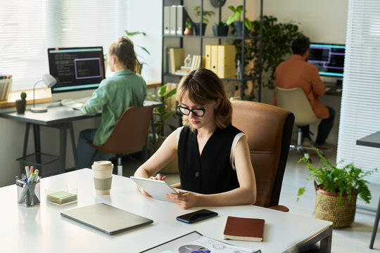 Caucasian young adult woman using digital tablet at desk, working on neural network development while two colleagues, Caucasian young adult man and woman, coding on computers in modern office