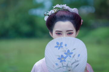 Traditional Japanese  pretty woman in Furisode Kimono holding fan and posting in green natural Pine Forest. Cultural fashion and nature harmony.