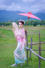 Traditional Japanese Woman in Furisode Kimono with red wagasa umbrella, fan, in a peaceful natural green. Cultural fashion and nature harmony.