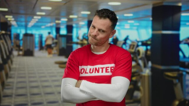 Young man wearing volunteer shirt making funny faces in gym with exercise equipment in background, showcasing humorous expression in an active setting.